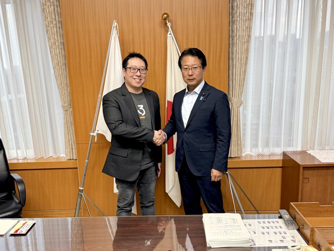 Two men shake hands in a formal office setting, with Japanese flags visible in the background, including a document on the table featuring numeric "3" markings. The image appears to depict a meeting between a Bitcoin magazine representative (likely the man in the "3" graphic tee) and a Japanese government official, relating to the contextual post about advocacy efforts to persuade state leaders to adopt Bitcoin reserves.