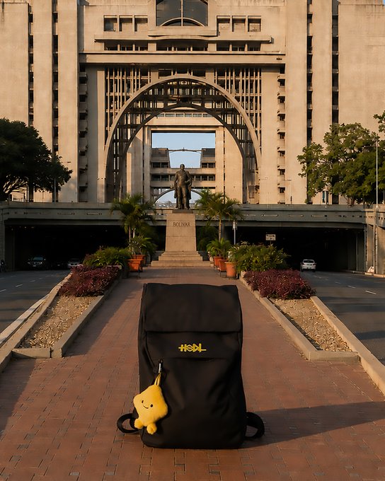 A black backpack with the yellow "HODL" logo and a small plush charm sits on a brick walkway in front of a grand architectural building featuring a large archway and a central statue. The image aligns with the post by showcasing the "HODL" branded backpack, symbolizing the traveler's enthusiasm for visiting Venezuela while embodying the crypto community's motto.