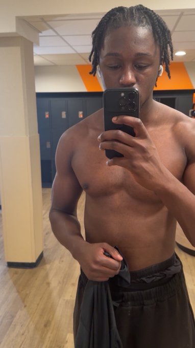 A shirtless young man with braided hair takes a mirror selfie in a gym locker room, holding a dark shirt.
