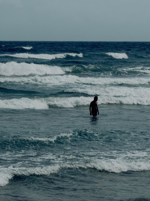 A lone figure wades into churning ocean waves under an overcast sky, capturing the solitary, anticipatory mood of waiting for AVAX to reach its all-time high this summer.