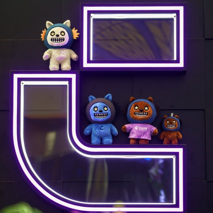 Four plush toys with star-eyed, grinning faces sit on illuminated purple LED shelves against a dark wall, with one wearing a shirt that reads "coinw."
