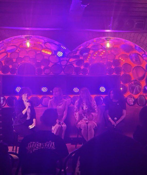 A dimly lit venue bathed in purple light, showing four people sitting on stools on a stage in front of a circular-patterned backdrop, with audience members visible in the foreground.