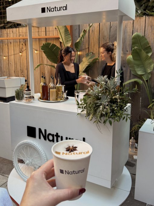 A person holds a coffee cup branded 'Natural' in front of a matching branded cart staffed by two women, decorated with plants and bottles of syrup.