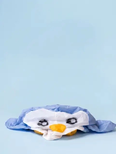 A crumpled tissue paper craft shaped like a blue penguin character with white face details and an orange beak rests on a light blue surface.