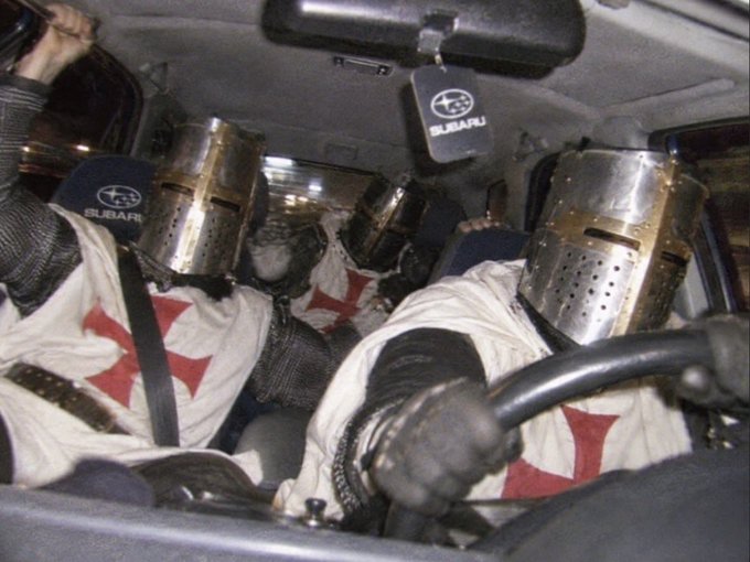 Four medieval knights in chainmail and helmets emblazoned with red crosses cram into a Subaru, wearing seatbelts and gripping the steering wheel, as if preparing to charge into battle. The visible "SUBARU" logos on their headrests and gear shift tag humorously reinforce the caption "gm, let's win" by blending ancient crusader imagery with modern suburban readiness.