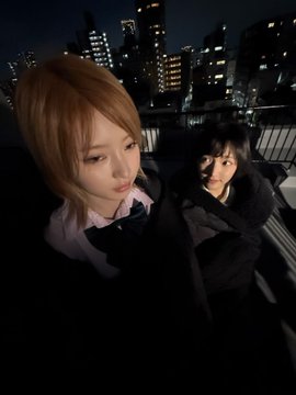 Two people pose closely together for a nighttime photo on what appears to be a rooftop or elevated area, with the illuminated city skyline visible in the background. The person in the foreground has light brown hair and wears a white shirt with a dark bow, while the person behind has dark hair and wears a dark jacket.