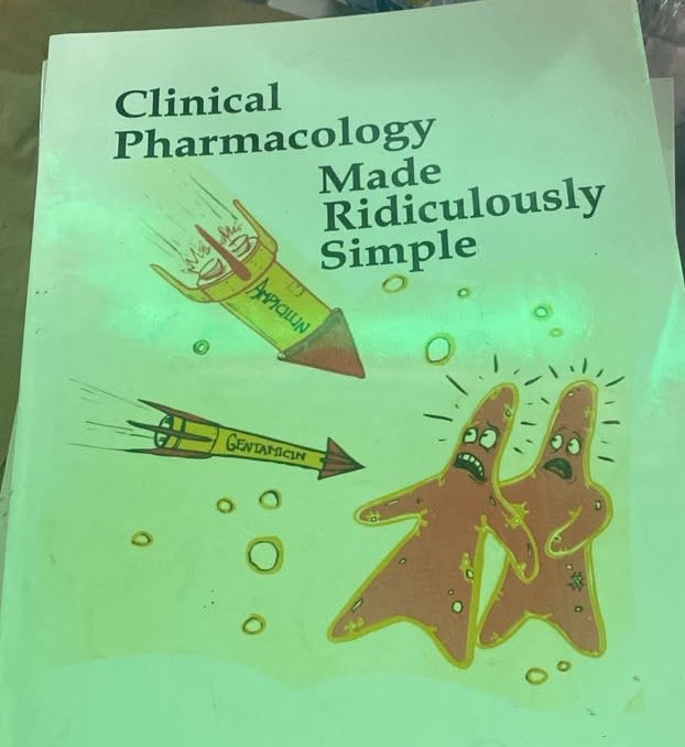 A worn copy of the medical book "Clinical Pharmacology Made Ridiculously Simple" sits on a desk, its cover featuring cartoonish illustrations of Ampicillin and Gentamicin capsules flying like rockets toward frightened, anthropomorphic brown starfish characters amid green-tinted bubbles. The accompanying post caption "𝐒𝐜𝐡𝐨𝐨𝐥 𝐢𝐬 𝐭𝐫𝐞𝐬𝐬𝐟𝐮𝐥" humorously reflects on the overwhelming and intense nature of studying pharmacology, suggesting that even powerful antibiotic "weapons" like Ampicillin and Gentamicin are needed to combat the stress and anxiety that medical or pharmacy students face while tackling this notoriously dense subject material.