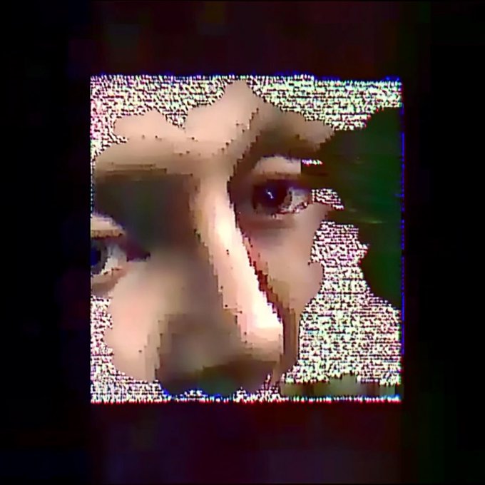 A heavily distorted, glitch-effect close-up of a person's face in black and white with static noise, displayed within a square frame against a dark background.