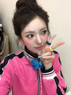 A young woman with her hair in a high bun wears a pink, black, and white track jacket with blue headphones around her neck, flashing a peace sign while displaying colorful rings on her fingers, aligning with the playful "#Sui Bubble" theme.