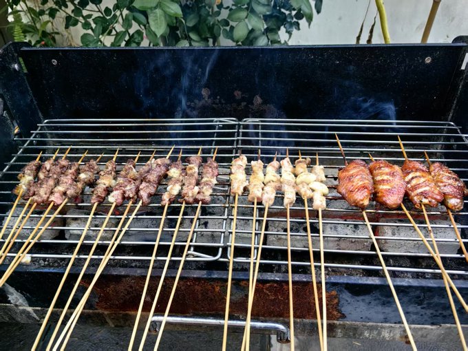 A black charcoal grill smokes with rows of meat skewers, including beef and chicken, cooking over the heat.