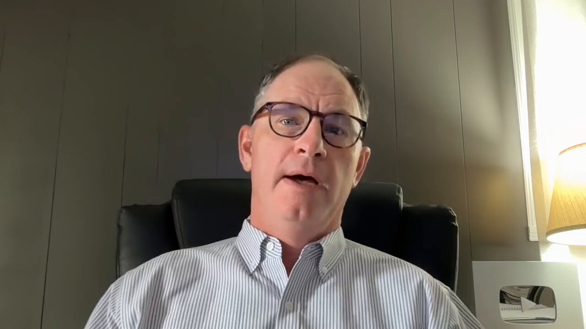 The image shows a man wearing glasses sitting in an office chair, looking at the camera with a serious expression. He appears to be engaged in a video call or participating in a live streaming event. The background of the image features a gray wall, which contrasts with the white lamp that is visible behind him. The text "XRP Price Before Law Watch" can also be seen in the image.