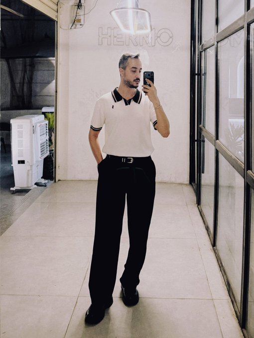 A man takes a mirror selfie while wearing a white polo shirt with black trim and black trousers, secured with a black belt featuring a silver buckle.