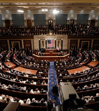 The image shows the U.S. Senate chamber filled with members seated at their desks, with the Speaker's rostrum and American flag centered at the front. A television camera in the foreground broadcasts the session, providing a fitting backdrop to the news that Citi bank is urging the Senate to pass the Crypto Clarity Act.