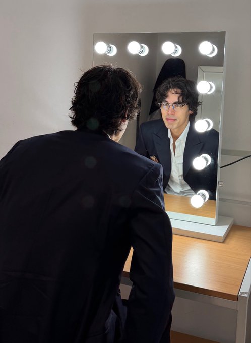 A man with curly dark hair and glasses wearing a dark suit jacket and white shirt looks at himself in a vanity mirror framed with bright round bulbs.