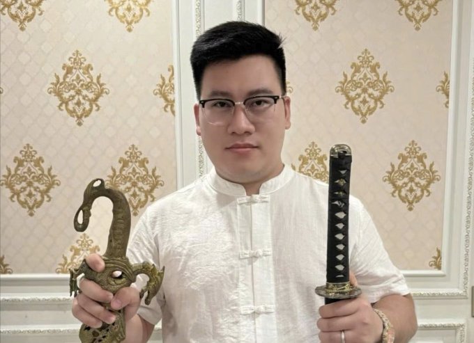 A man wearing glasses and a white traditional Chinese shirt holds a bronze dragon-shaped decorative piece in one hand and a black-handled katana in the other.