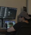 A person wearing a cap sits in a dark room illuminated by multiple computer monitors displaying code and data.