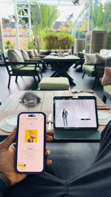 A person holding a smartphone displaying an NFT gallery while sitting in an outdoor cafe or lounge area with a tablet visible on the table beside them.