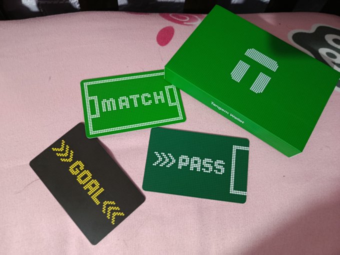 A green Tangem Wallet box sits alongside three pixelated-style cards labeled ">>match", ">>GOAL", and ">>PASS," all resting on a pink surface. The image ties to the post by showcasing the collaboration's playful take on a soccer/football victory theme with the "GOAL" and "match" cards celebrating a win alongside the Tangem crypto wallet packaging.