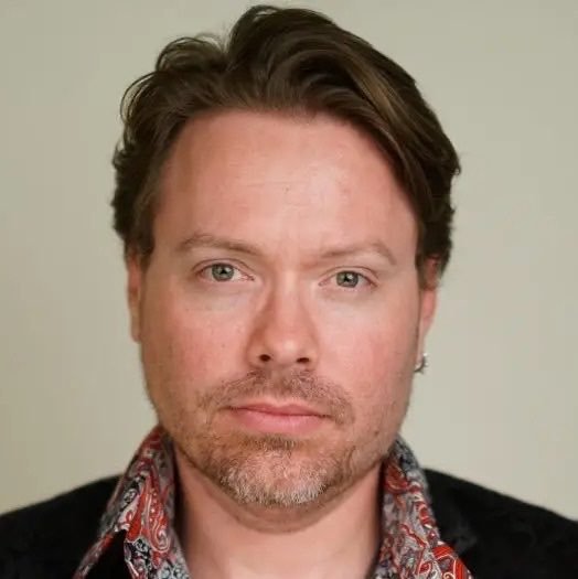 Headshot of a man with light brown hair and a short beard wearing a patterned scarf against a neutral background.