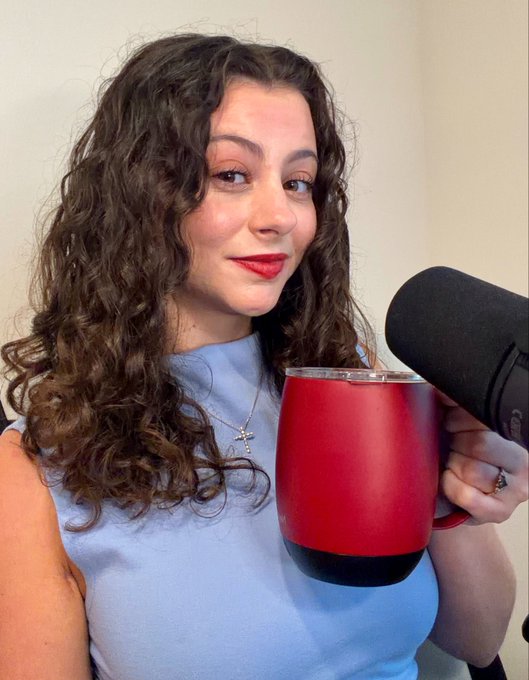 A woman with curly brown hair and red lipstick holds a red tumbler while smiling next to a podcast microphone, promoting her upcoming appearances on Newsmax to discuss the Clarity Act and Bitcoin, followed by Daily Zest.