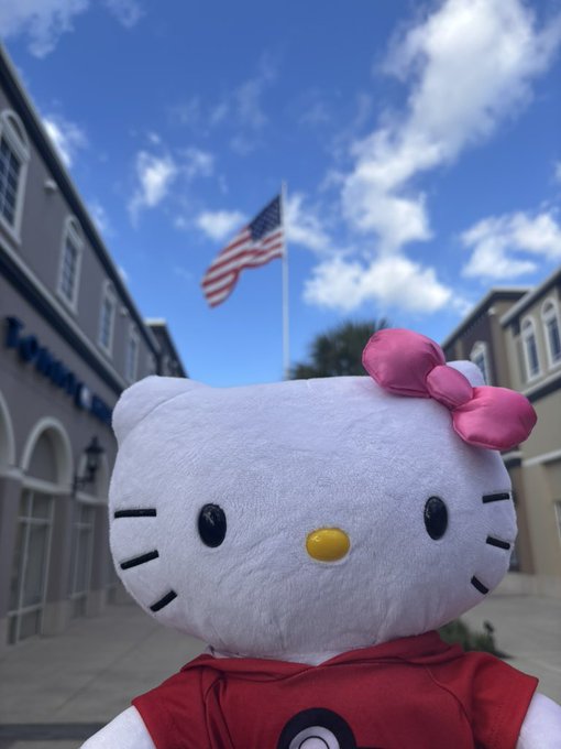 A Hello Kitty plush toy wearing a red outfit and pink bow is held up against a backdrop of a blue sky with clouds, an American flag, and buildings.
