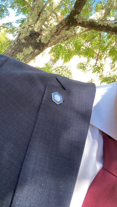 A close-up of a dark grey suit jacket lapel pin features a small blue hexagonal design, worn with a white shirt and burgundy tie outdoors.