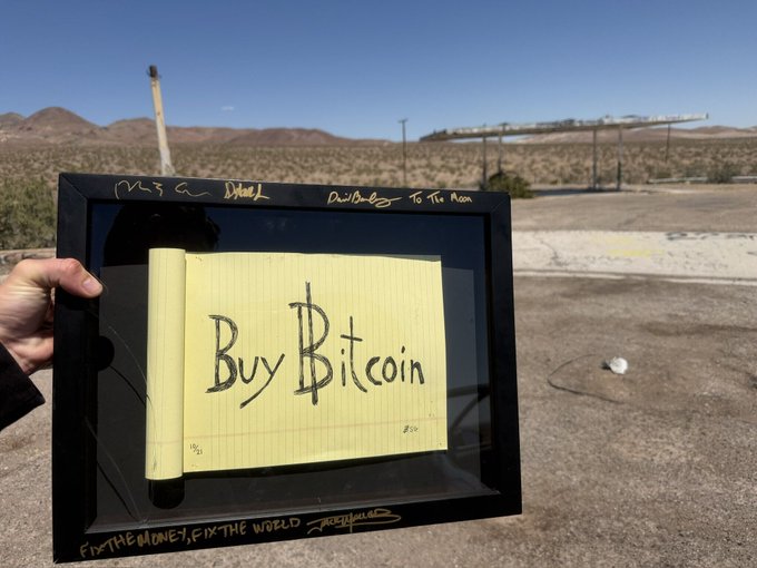 A framed piece of yellow legal pad paper with "Buy Bitcoin" handwritten in black marker, signed by FreeSpeechMD with the phrase "FIX THE MONEY, FIX THE WORLD," held in a desert landscape.