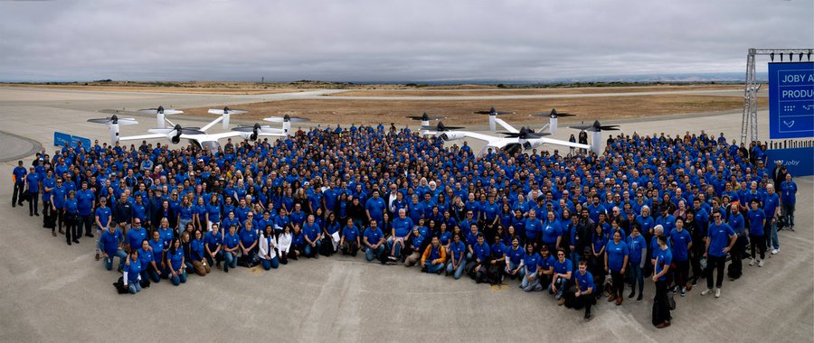 Hundreds of Joby Aviation employees in matching blue shirts gather for a group photo on an airfield, posed in front of several white electric vertical takeoff and landing (eVTOL) aircraft prototypes, with a banner on the right displaying "JOBY A[VIATION] PRODU[CTION]" and the ticker "$JOBY," visually reinforcing the post's message about leadership driving the company's progress.