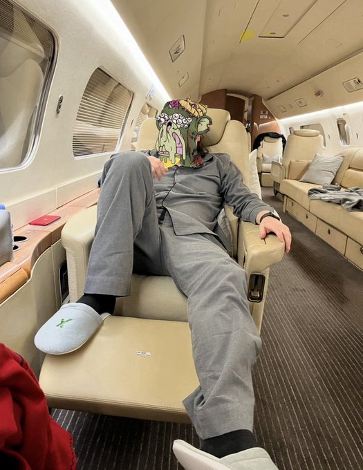 A person wearing a cartoonish mask and gray loungewear reclines in a plush private jet cabin, wearing white Hype slippers with black socks, embodying the "gn" (good night) vibe of luxurious travel comfort before sleep.