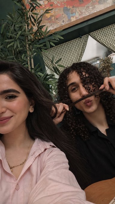 Two young women pose together for a selfie, with one in a pink shirt and the other with curly hair holding what appears to be a guitar, in front of plants and artwork.