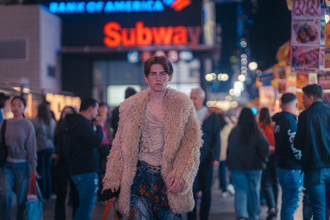A young man with red hair wearing a fuzzy tan coat stands on a city street at night, with a 'Subway' sign and 'Bank of America' sign glowing in the background.