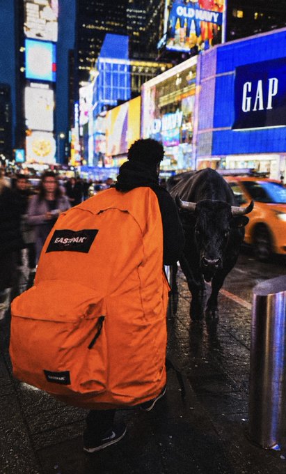 A street photograph taken in Times Square, NYC at night showing a person wearing a large orange Eastpak backpack standing next to a bull statue with the GAP store sign visible in the background.