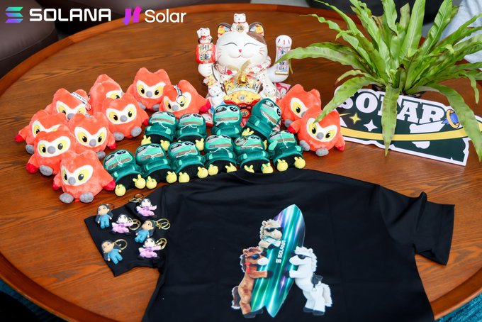 A display of Solana/Solar merchandise on a wooden table, including orange fox plushies, green turtle plushies, a black t-shirt featuring axolotls on a surfboard, a cat figurine, keychains, and a Solar flag, with Solana and Solar logos at the top.