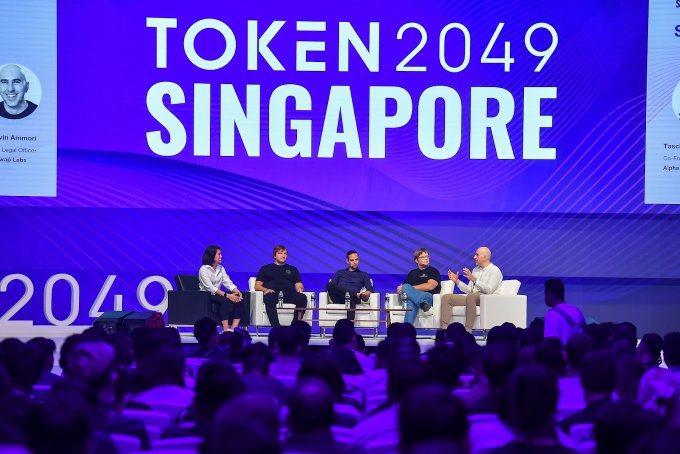 A panel discussion at TOKEN 2049 SINGAPORE, with five speakers seated on stage in front of a large screen displaying the event name, while an audience watches from below.