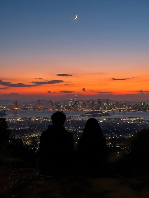 A silhouette of a couple sits together on a hillside, gazing out at the San Francisco skyline illuminated beneath a crescent moon and a vibrant sunset sky. The serene view captures a quiet moment of stillness, reflecting the post's theme of "Peace."