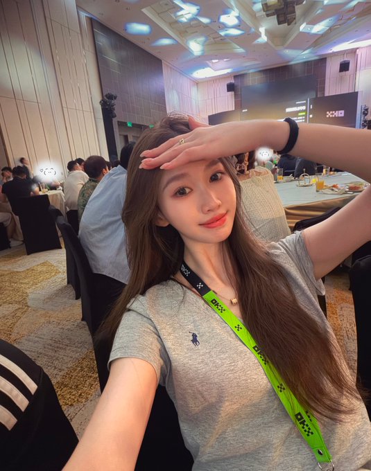 The image shows a young woman taking a selfie at what appears to be a corporate event, wearing a gray Ralph Lauren t-shirt and a green OKX lanyard, with one arm raised to her forehead in a casual pose. Behind her, attendees can be seen seated at tables in a banquet hall setting, suggesting a networking dinner at the OKX Growth Accelerator event in Macau. The green OKX lanyard she's wearing and the professional conference setting align with the post's mention of meeting new friends on the first day of the OKX Growth Accelerator Macau stop.