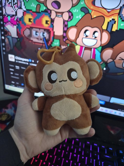 A hand holds up a brown cartoon-style monkey plush keychain in front of a computer monitor displaying colorful NFT artwork, with a pink and purple backlit keyboard visible below.