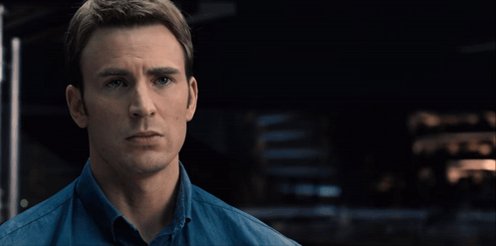 The image features a man wearing a blue shirt, looking at the camera with an intense expression. The background of the image shows a city street at night, which adds to the overall atmosphere of the scene. There are no texts or other objects present in the image, and the focus is solely on the man's face and his serious expression as he looks directly into the camera.