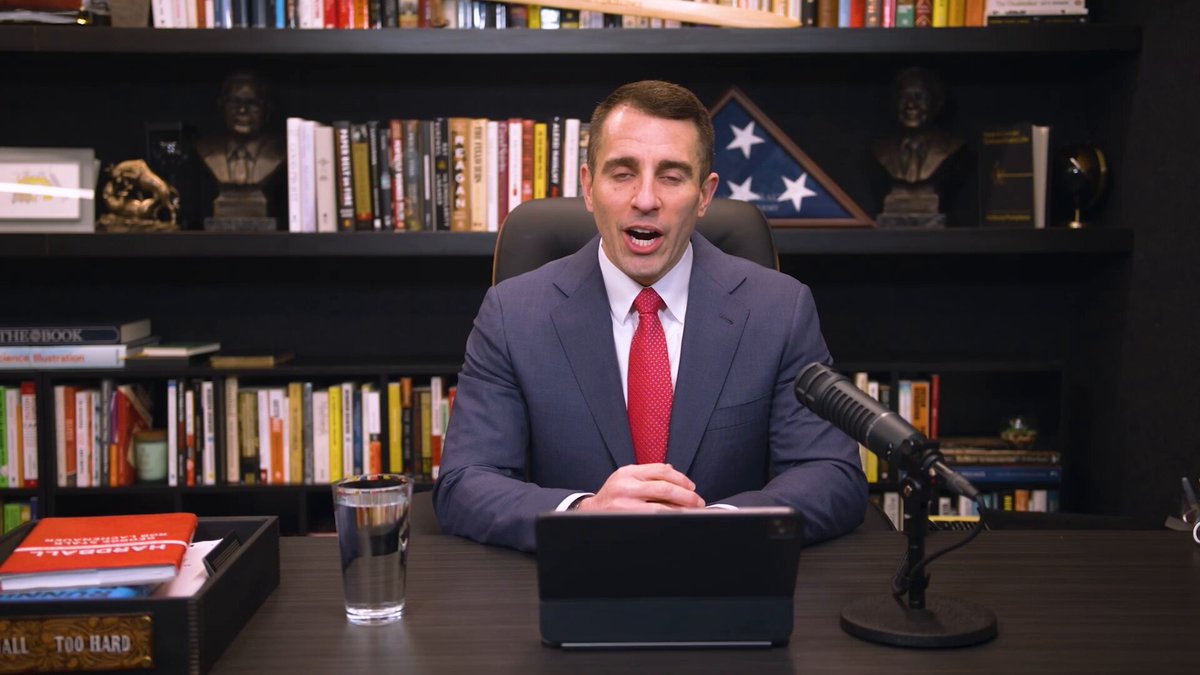 The image shows a man sitting at a desk with a laptop, microphone, and glass of water. He appears to be wearing a suit and tie, suggesting that he might be a professional or an important figure. The text "Bitcoin is crashing. Stocks are up." indicates that there has been a significant increase in Bitcoin's value recently, which could have caught the attention of the man sitting at his desk.