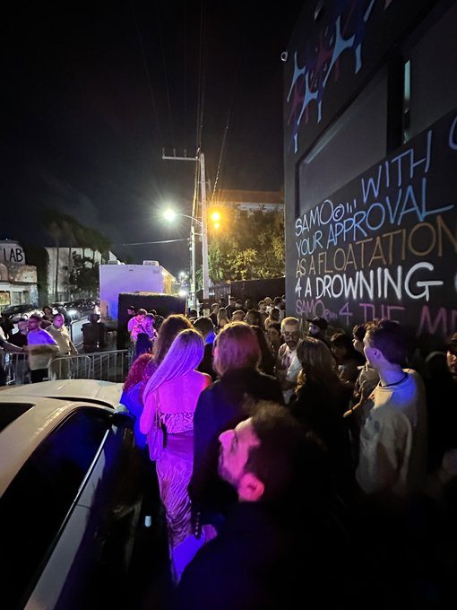 A nighttime photograph of a crowd gathered outside a venue, with purple lighting on the people and a wall displaying the text "SAMOON WITH YOUR APPROVAL AS A FLOATATION A DROWNING".