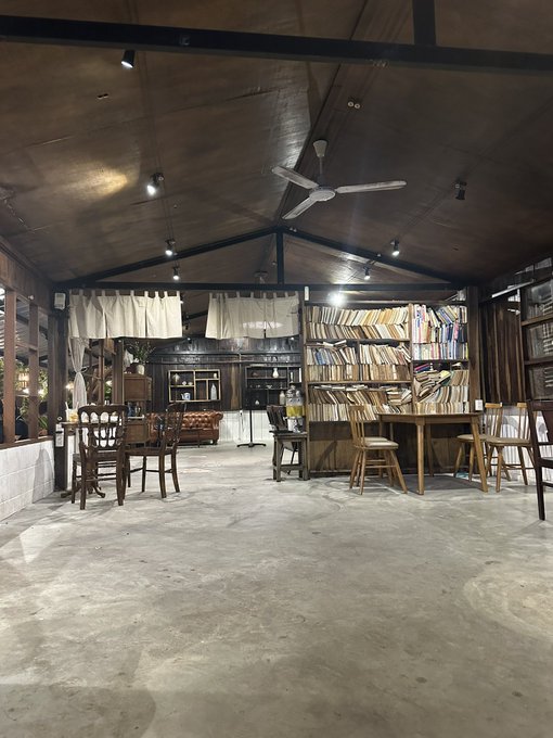 An empty café interior features a high wooden ceiling with a ceiling fan, wooden tables and chairs, a leather sofa, and tall bookshelves filled with books.