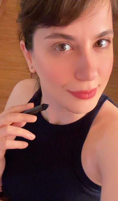 Close-up selfie of a young woman with short brown hair wearing red lipstick and a black top, holding a small black object near her face.