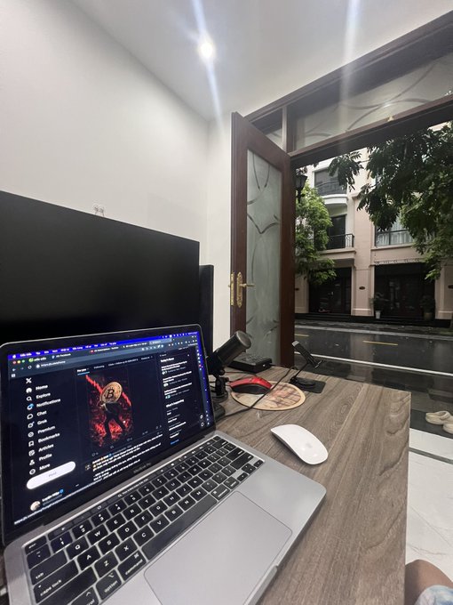 The image shows a laptop computer sitting open on top of a wooden desk, with an empty mousepad next to it. The background features a window overlooking a street outside. The photo appears to be taken from behind the laptop screen, giving a unique perspective on the scene. The content of the post is not clear or identifiable, but the image seems to be related to Bitcoin and cryptocurrency discussions.
