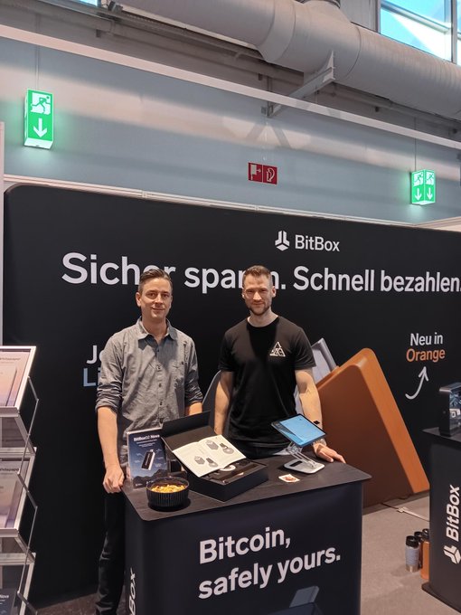Two men stand at the "Bitcoin, safely yours." XAUUSD/s (Gold vs US Dollar / Spot) Bitcoin booth, though the post indicates the Frankfurt team (Markus) and Helsinki team (Pako and Laura) are spread across two events—Finance26 and BTCHEL.