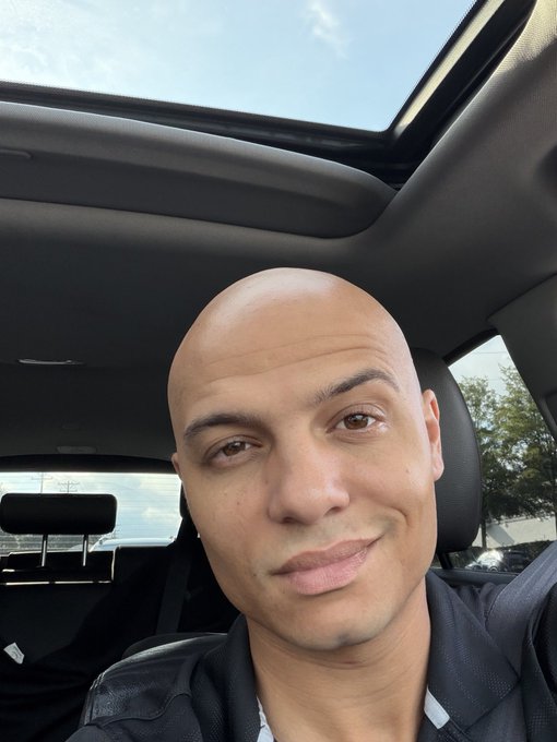 A selfie of a bald man smiling inside a car with a sunroof open to a blue sky.