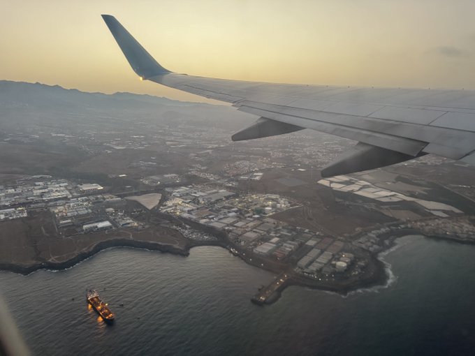An airplane wing is visible in the foreground as the plane departs Gran Canaria, overlooking the island's coast and a cargo ship. This image complements the post about leaving the island and returning to work on a crypto market report newsletter.