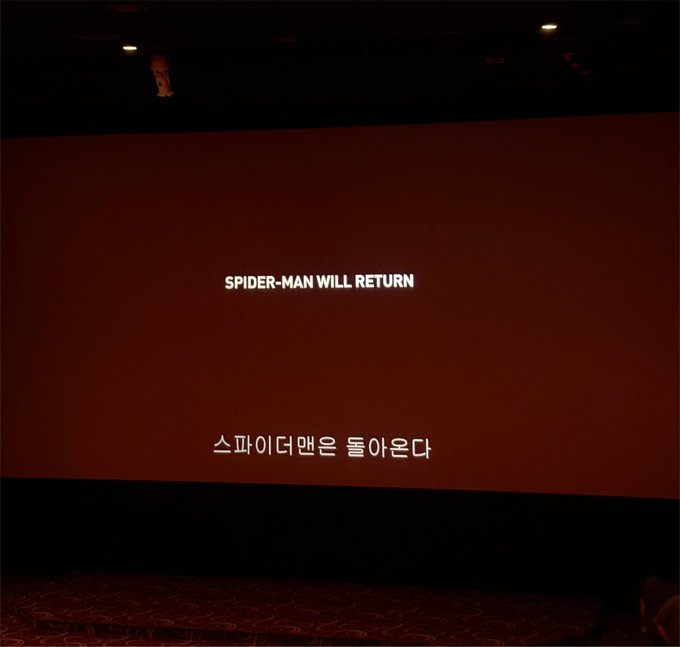 A movie theater screen displays a post-credits scene teaser with the text "SPIDER-MAN WILL RETURN" in English and its Korean translation "스파이더맨은 돌아온다" below it. Paired with the caption "The crypto bull run is coming back," the image humorously likens the highly anticipated return of Spider-Man to the market's eager anticipation of a new crypto bull run, using the iconic Marvel cliffhanger format as a playful parallel.