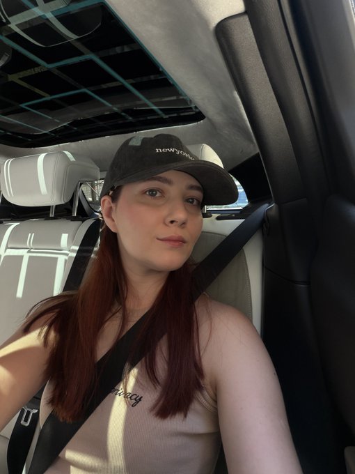The image features a woman wearing a black baseball cap, sitting inside a car. She has long hair that falls over her shoulders as she looks out of the window. The photo captures her smiling face, suggesting that she is enjoying herself or having an interesting experience.