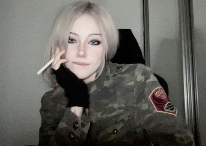 A young woman with blonde hair holds a cigarette while wearing a camouflage jacket featuring a red and gold military-style patch on the upper left arm.