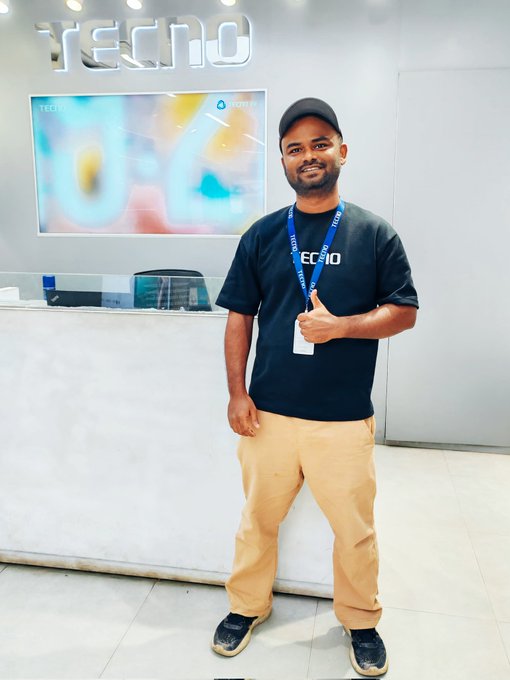 The image features a man standing next to a desk at a store or office, wearing a black shirt and khaki pants with a blue lanyard around his neck. The man appears to be smiling for the camera while posing near the counter. In the background of the photo, there is a TV screen mounted on the wall that displays information about Web3 projects. This suggests that the store or office may be related to technology or cryptocurrency, and the man could potentially be an employee or customer interested in these topics.